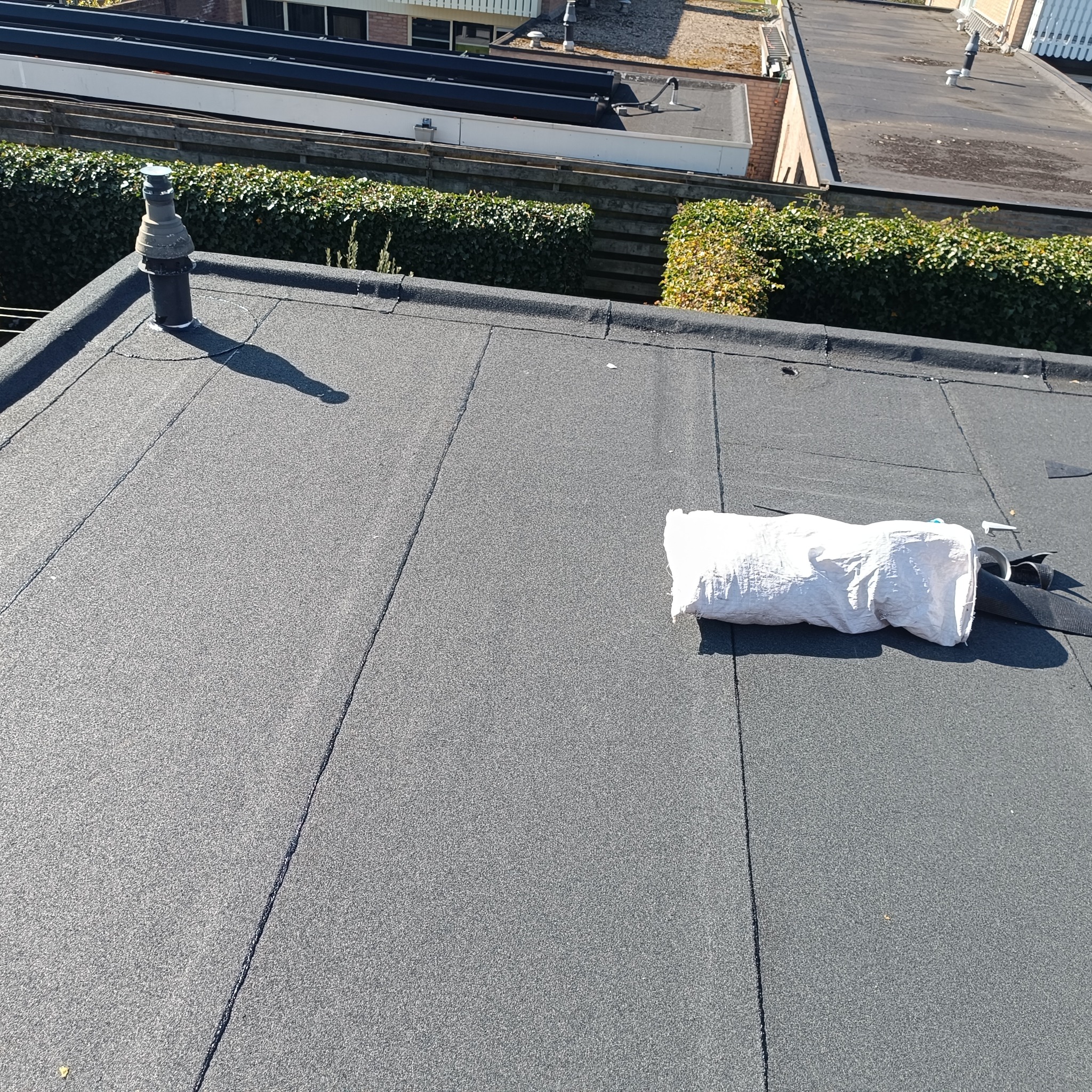 Czarny, płaski dach z widoczną wentylacją i workiem z narzędziami. Dach pokryty papą, na tle zielony żywopłot i budynki.