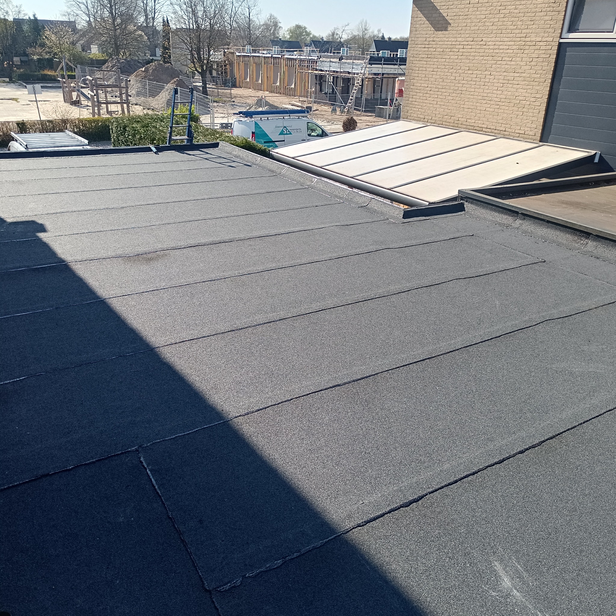 Szary, płaski dach pokryty papą termozgrzewalną, z widocznym cieniem i elementami konstrukcyjnymi w tle. W tle budowa i plac zabaw.