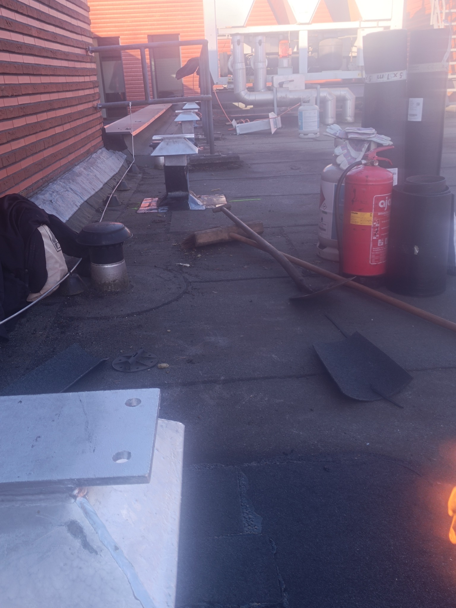 Prace na dachu z papy: narzędzia, wentylacja, gaśnica i rolki papy. Fragment muru z cegły w tle. Widoczne ślady niedawnych prac dekarskich.