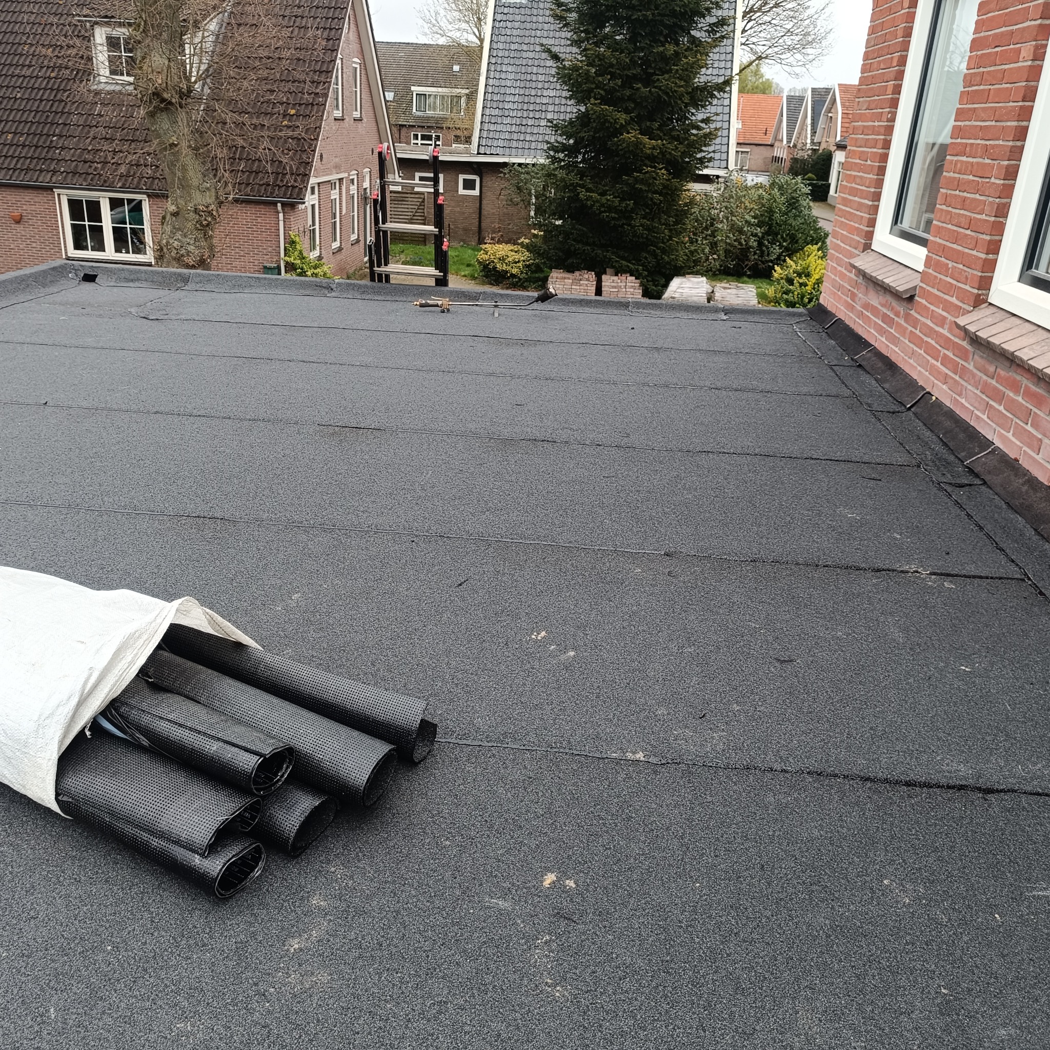 Gotowy płaski dach pokryty papą z widocznymi rolkami papy w worku. W tle zabudowa jednorodzinna, drabina oparta o budynek.