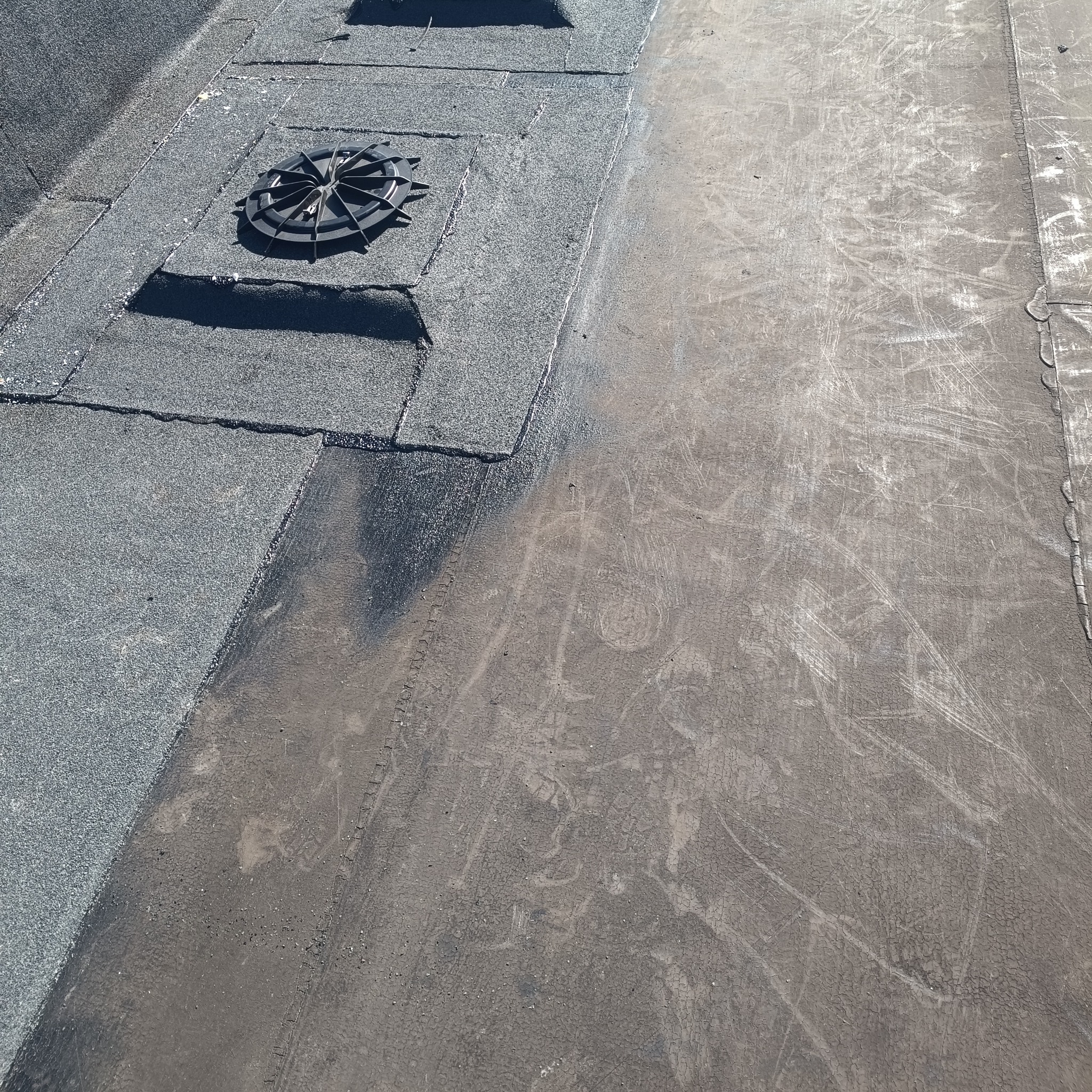 Fragment dachu z papy termozgrzewalnej, z widocznym wpustem dachowym i śladami napraw. Szara, matowa powierzchnia z wyraźną fakturą.