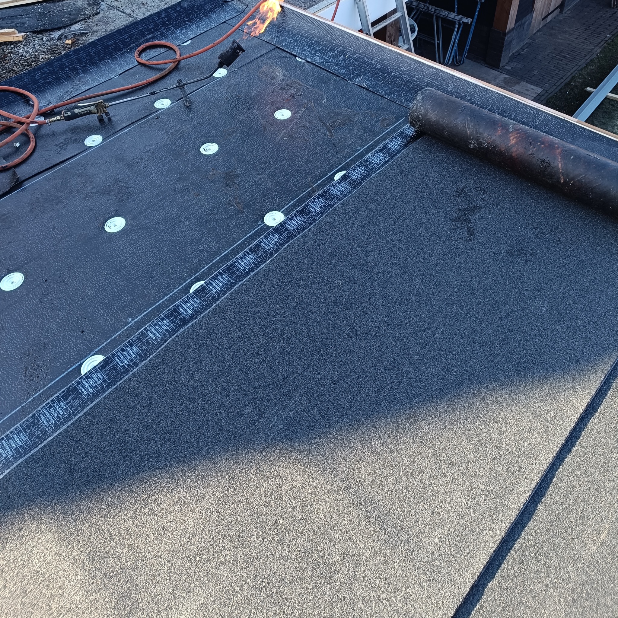 Krycie dachu papą termozgrzewalną z użyciem palnika w Wodzisławiu Śląskim. Widoczne mocowania i rolka papy na powierzchni dachu. Prace dekarskie w toku.