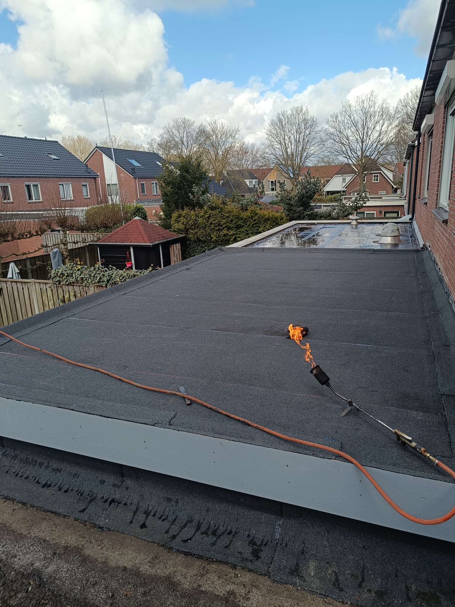 Czarny dach płaski z palnikiem w trakcie prac dekarskich. Widok na domy i drzewa w tle, błękitne niebo z chmurami. Pomarańczowy wąż od palnika leży na dachu.