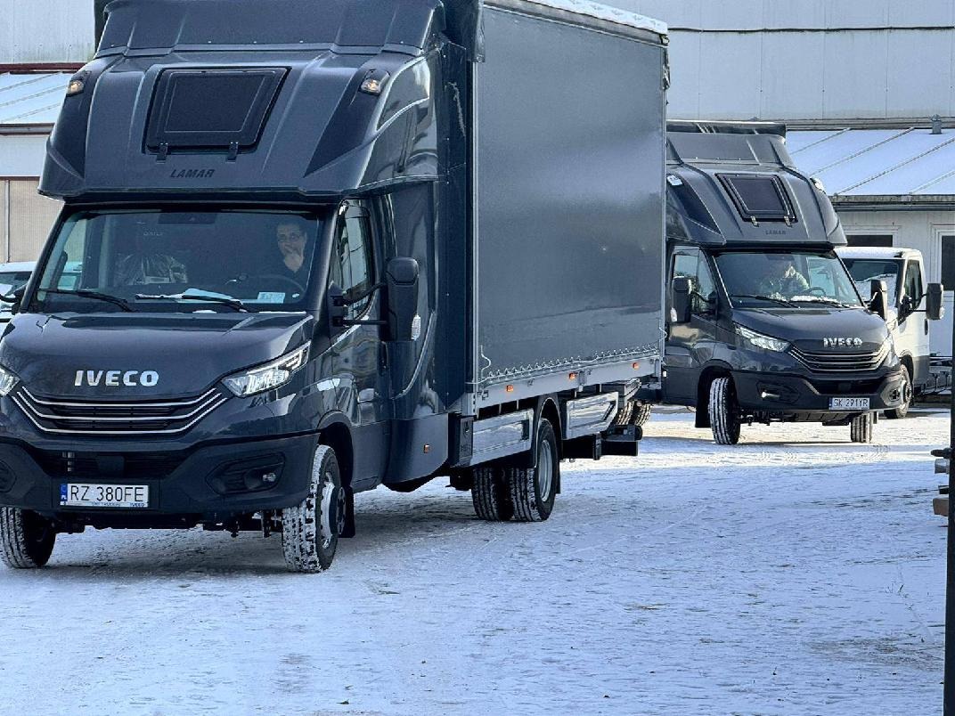 Dwa identyczne, ciemnoszare samochody ciężarowe marki Iveco z nadbudową LAMAR, zaparkowane na śniegu przed budynkiem w pochmurny dzień. Na pierwszym planie widoczny przód pojazdu z tablicą...