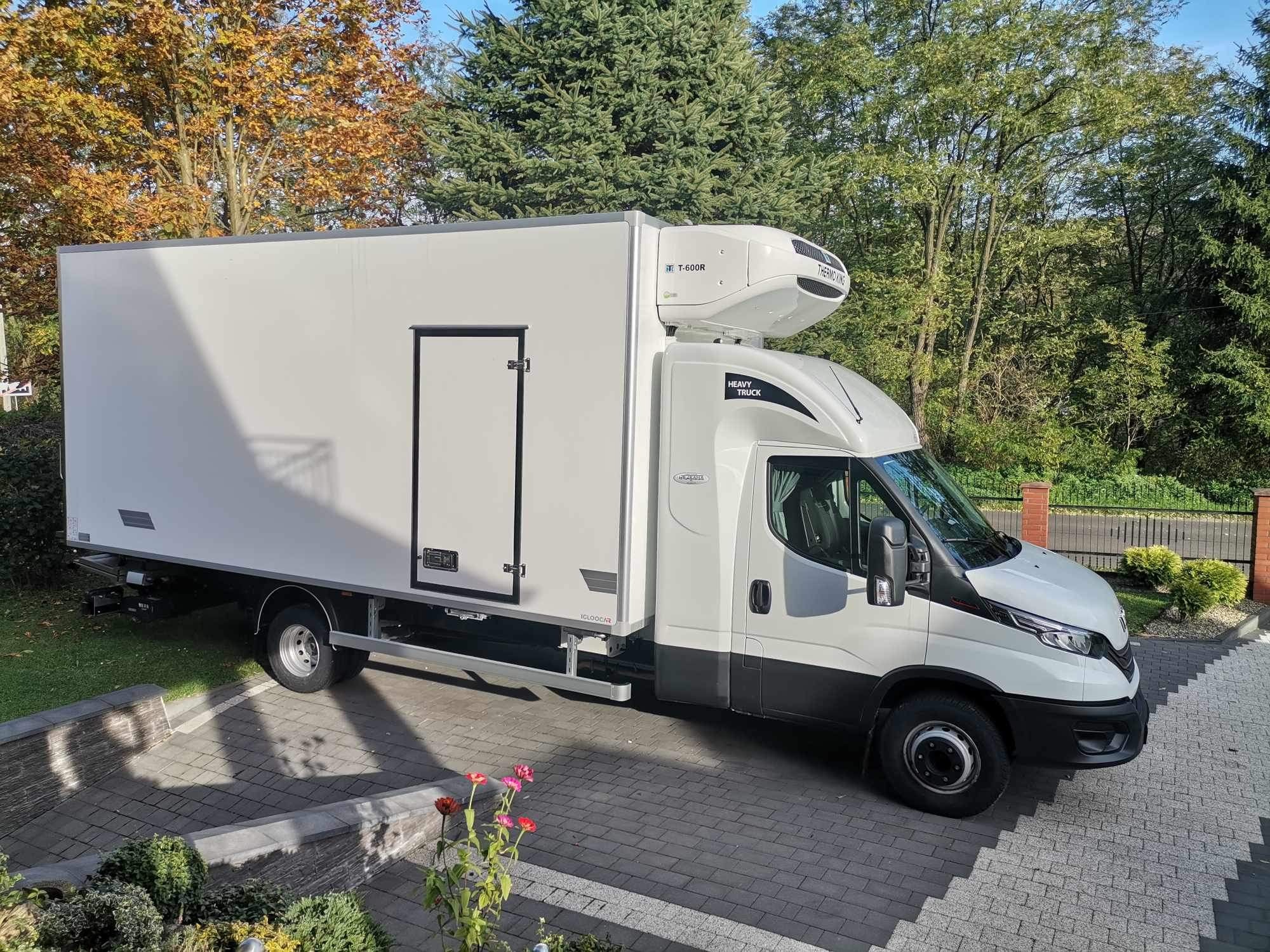 Biała ciężarówka chłodnia marki Iveco zaparkowana na brukowanym podjeździe, widoczne urządzenie chłodnicze Thermo King T-600R na dachu kabiny, w tle drzewa w jesiennych barwach.