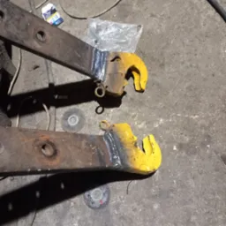 Dwa metalowe haki z przyspawanymi elementami, pomalowane na żółto, leżą na betonowej podłodze warsztatu obok zużytych tarcz do szlifierki i paczki papierosów.