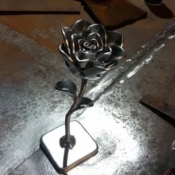 Ręcznie wykonana, metalowa róża z połyskliwego metalu na kwadratowej podstawie, widoczna na tle roboczego stołu z metalu, obok leżą rękawice spawalnicze i puszka farby w sprayu.