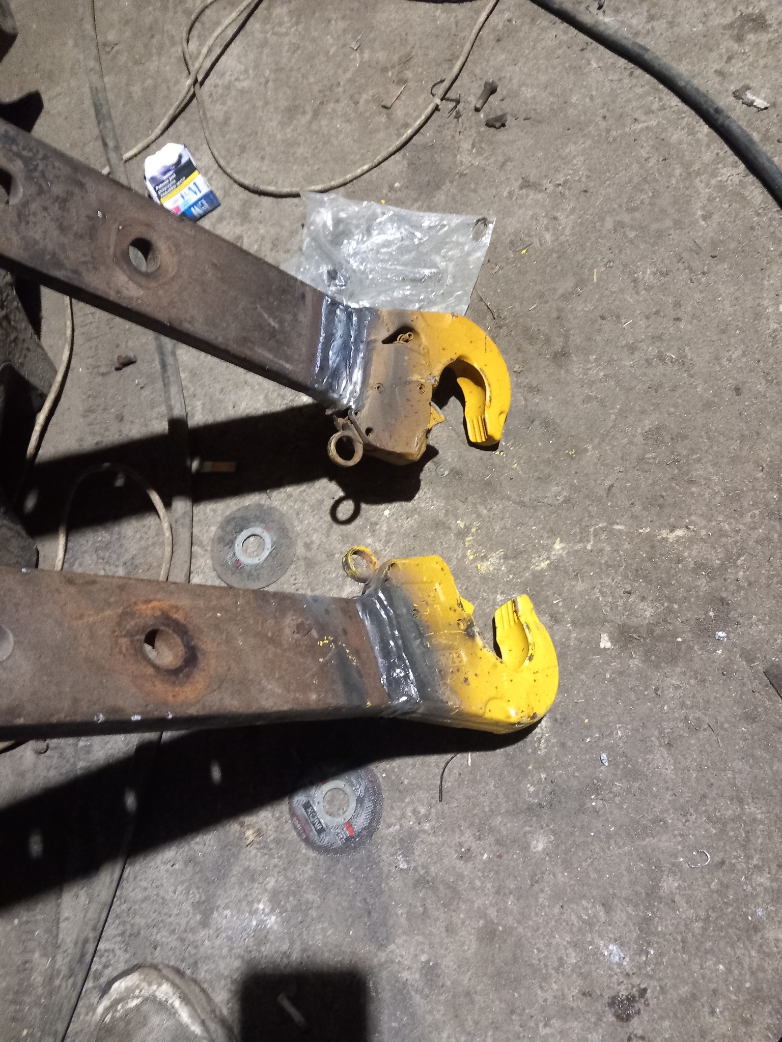 Dwa metalowe haki z przyspawanymi elementami, pomalowane na żółto, leżą na betonowej podłodze warsztatu obok zużytych tarcz do szlifierki i paczki papierosów.