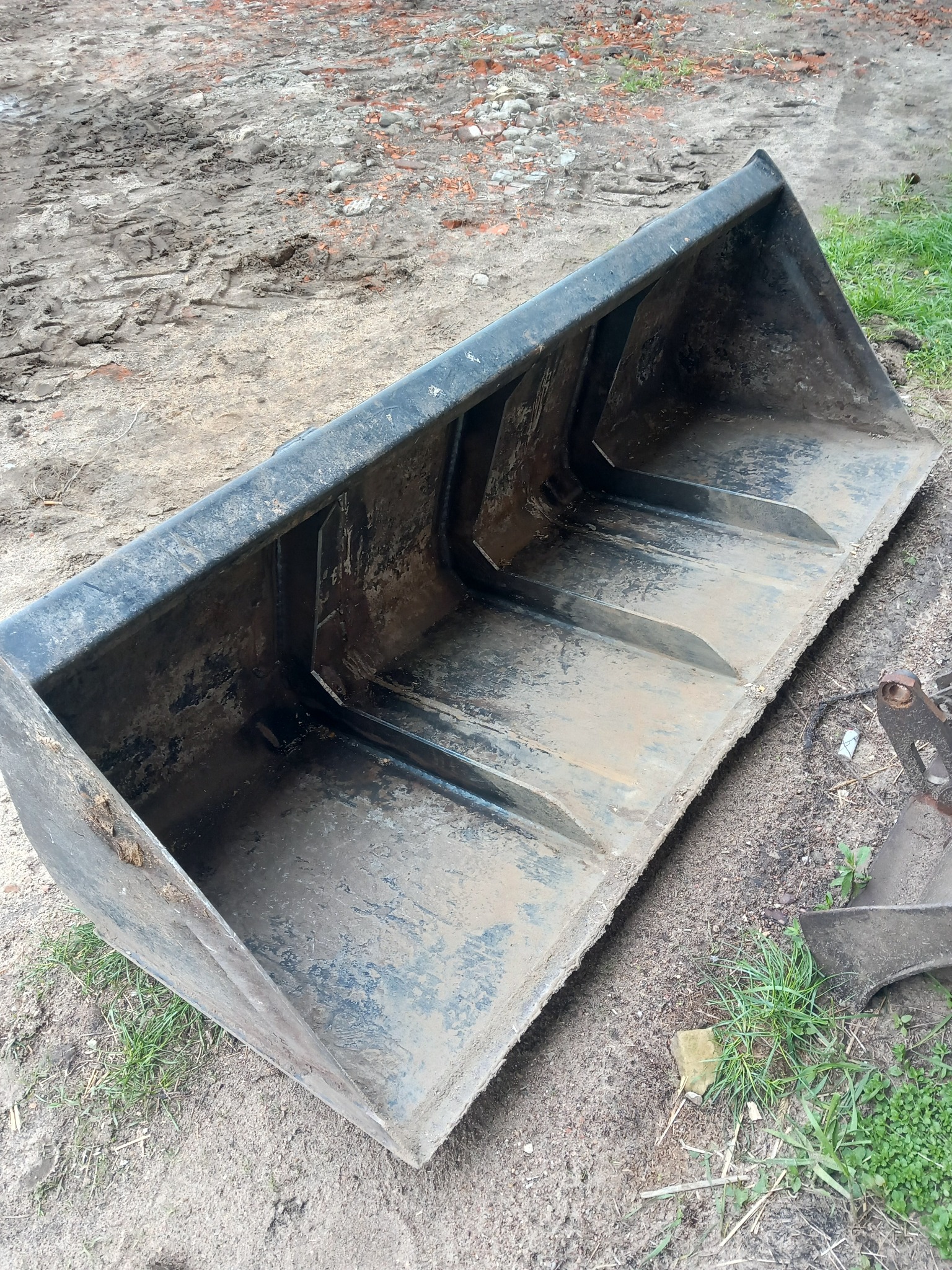 Używana, metalowa łyżka koparki leżąca na ziemi z błotem i trawą, widoczne ślady użytkowania i zabrudzenia.