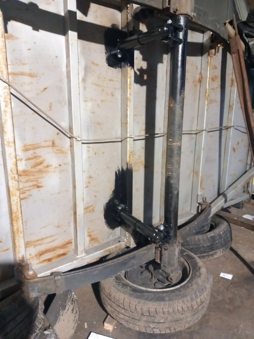 Spawana konstrukcja metalowa z osią i kołami, prawdopodobnie element bramy lub przyczepy, przymocowana do metalowej ściany z widocznymi śladami rdzy i spawania.