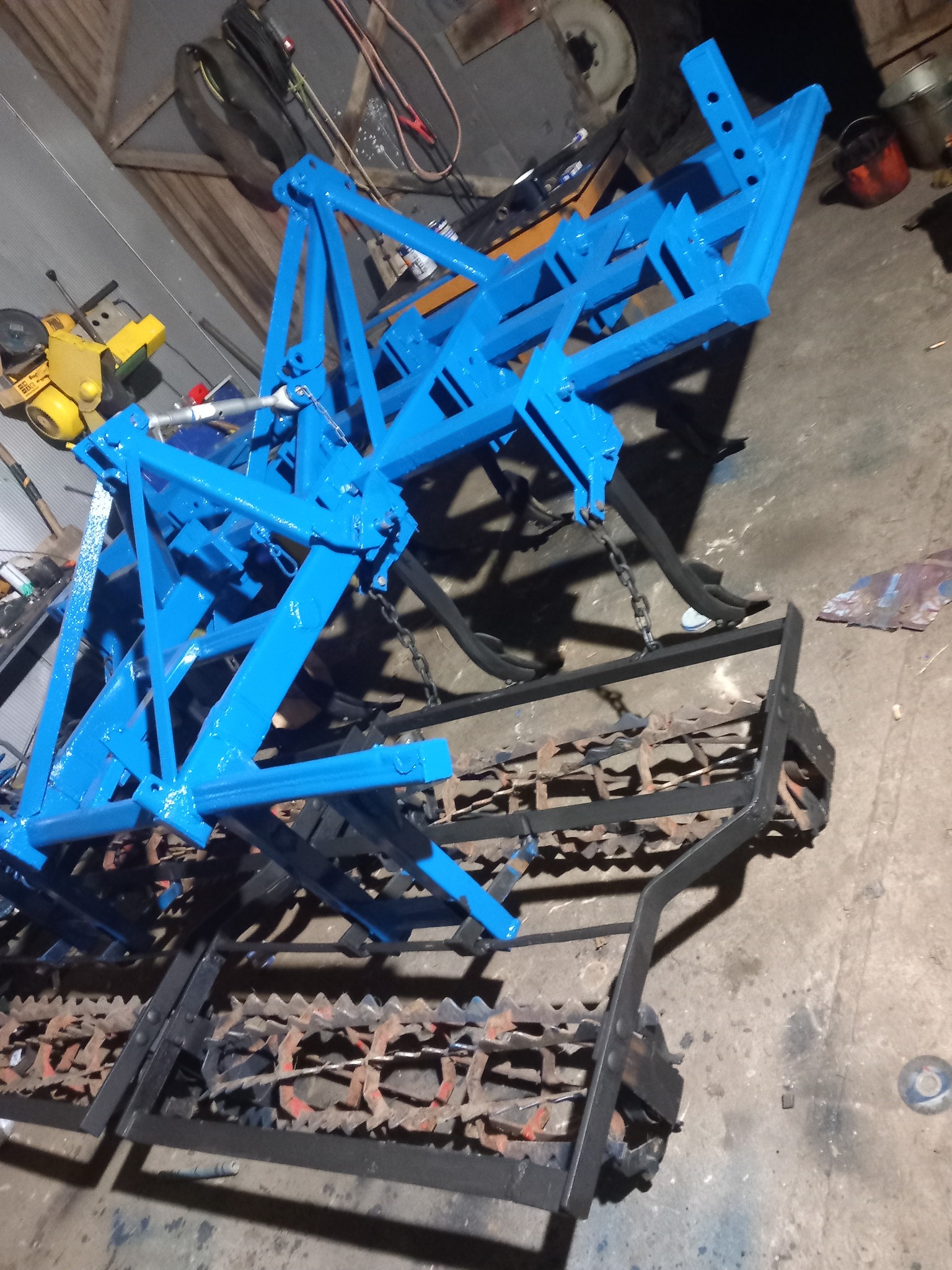 Niebiesko-czarna brona talerzowa, świeżo pomalowana, widoczna w warsztacie z narzędziami w tle.