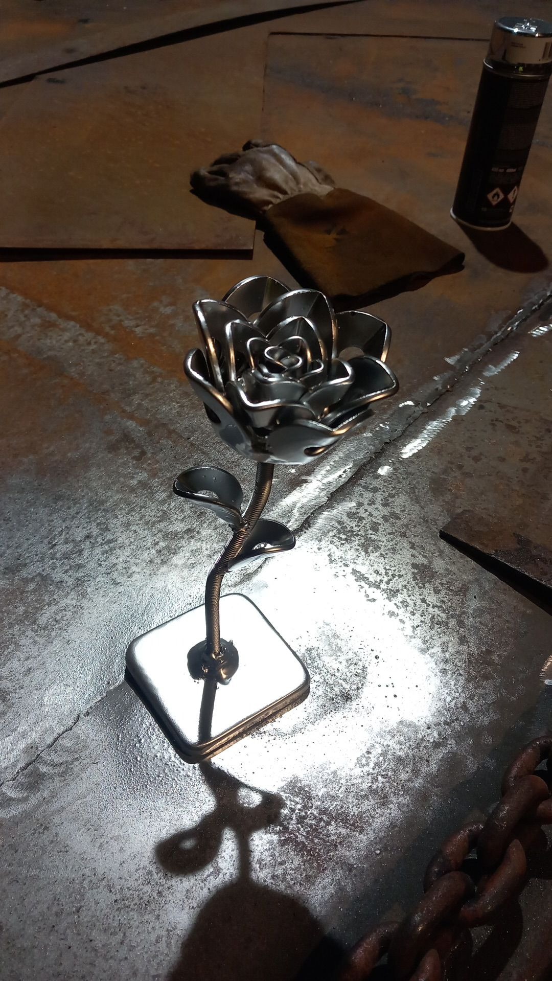 Ręcznie wykonana, metalowa róża z połyskliwego metalu na kwadratowej podstawie, widoczna na tle roboczego stołu z metalu, obok leżą rękawice spawalnicze i puszka farby w sprayu.