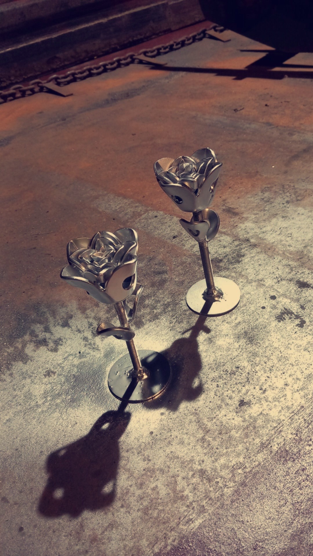 Dwie metalowe róże na metalowej powierzchni z łańcuchem w tle, wykonane z metalowych elementów i śrub, rzucające wyraźny cień.