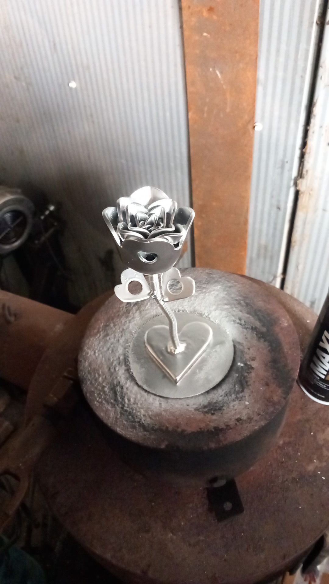 Ręcznie wykonana, metalowa róża z sercem na podstawie, stojąca na metalowym warsztacie, w tle blaszana ściana.