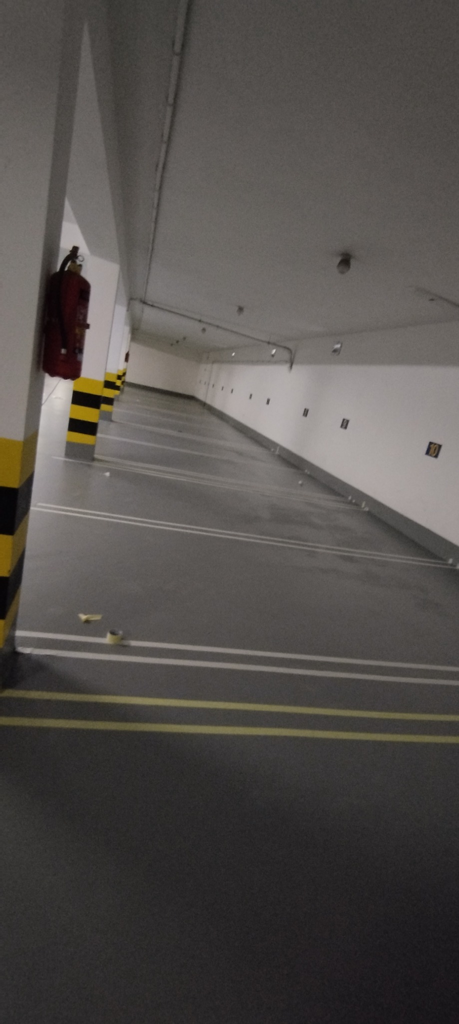 Pusta przestrzeń garażowa z pomalowanymi na szaro podłogami i białymi ścianami, słupami z czarno-żółtymi pasami i czerwonym gaśnicą na ścianie z lewej strony.