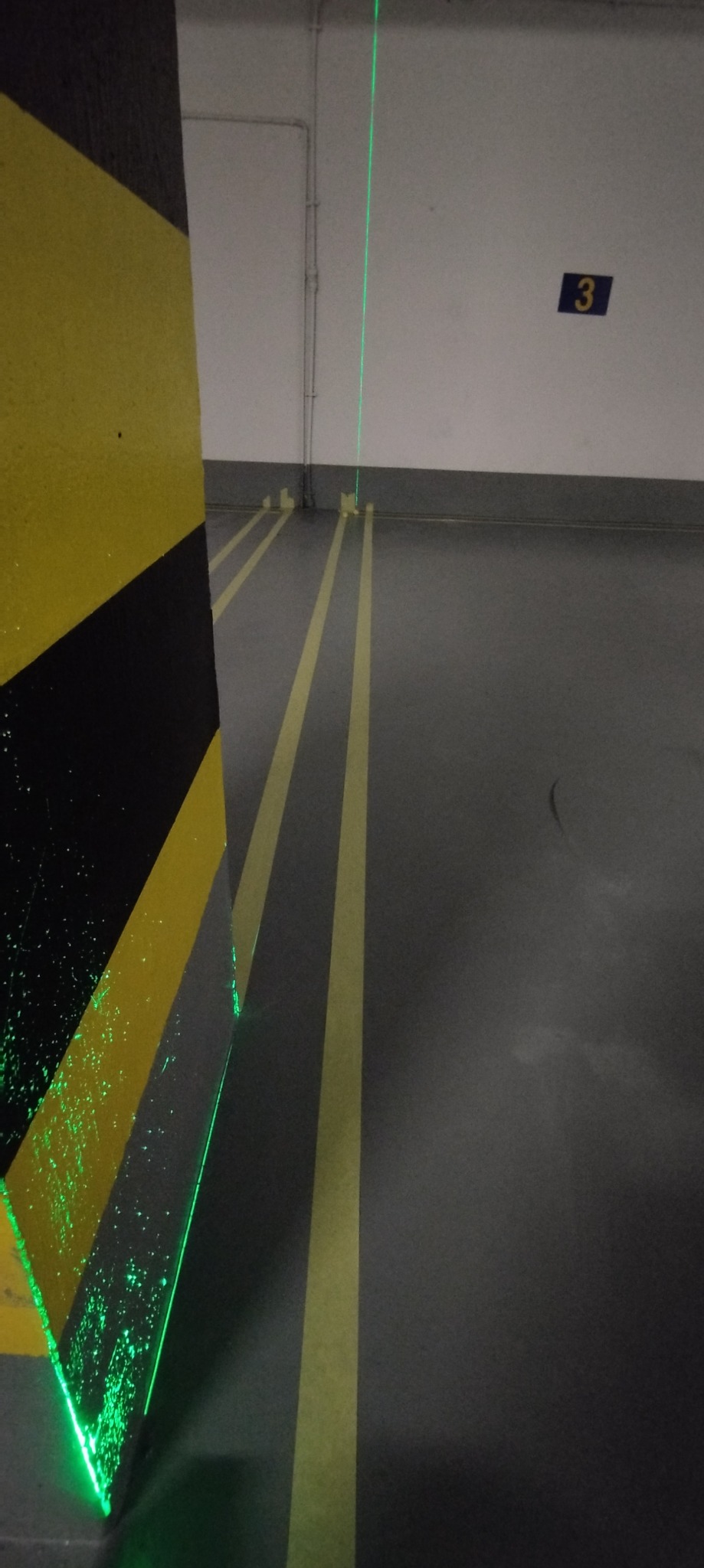 Laserowy niwelator pionowy wyświetla zieloną linię na ścianie i podłodze parkingu, wyznaczając pion i poziom w trakcie prac remontowych.