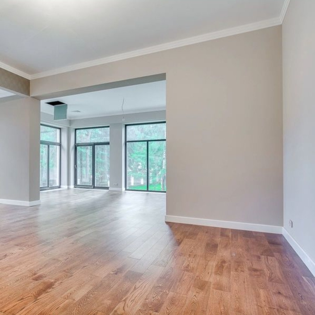 Jasne, puste wnętrze z drewnianą podłogą, szarymi ścianami i dużymi oknami z widokiem na zieleń na zewnątrz, listwy przypodłogowe w kolorze białym.