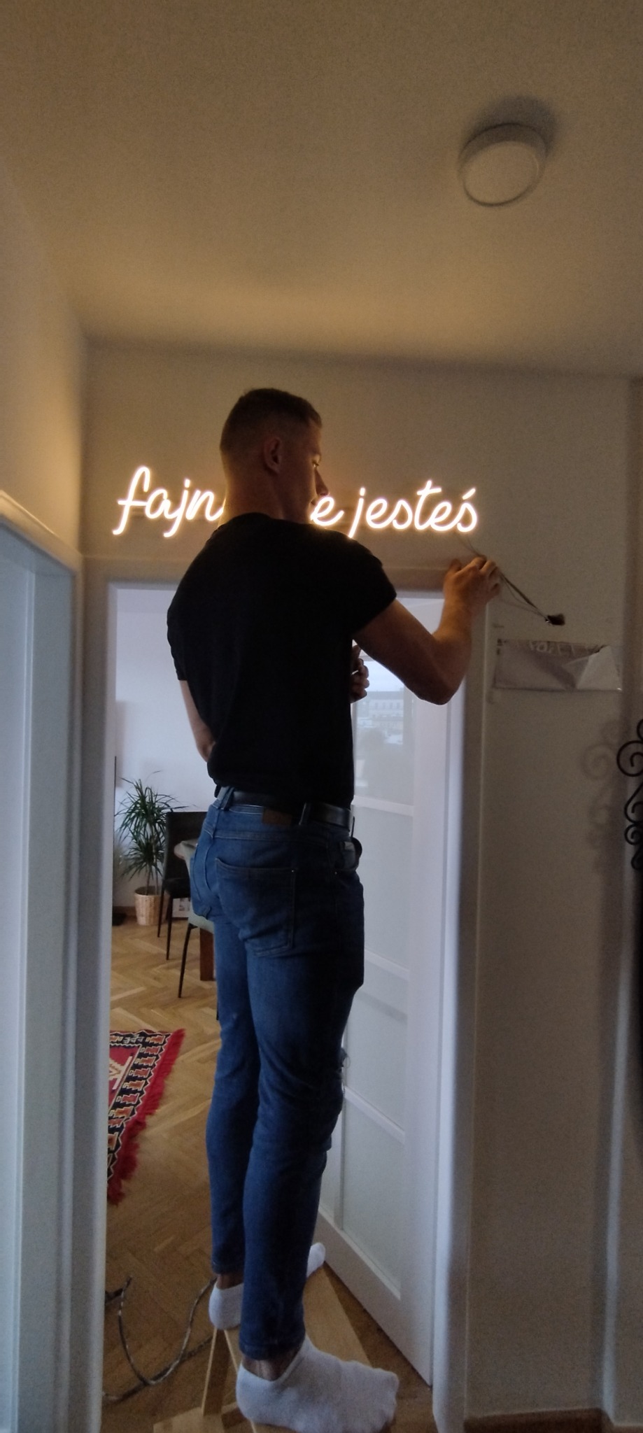 Mężczyzna na drabince montuje neonowy napis 'fajne jesteś' na ścianie w korytarzu, widoczne przewody i uchwyt montażowy.