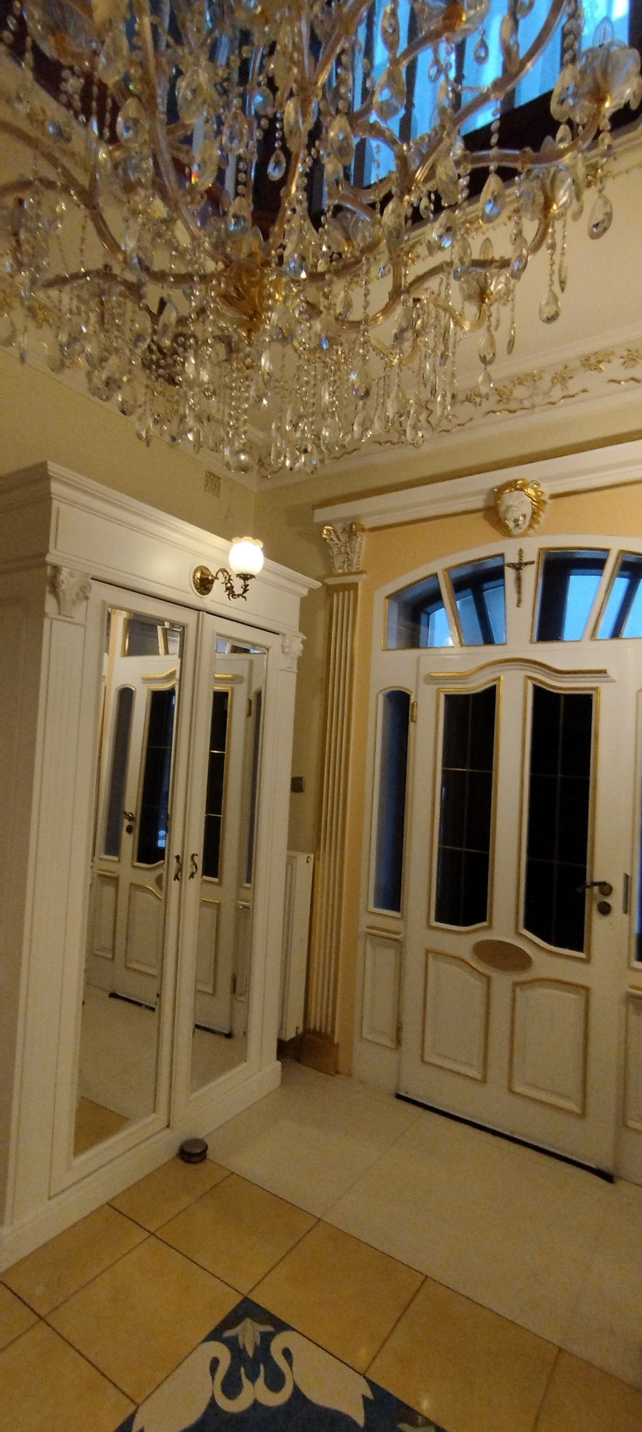 Elegancki przedpokój z kryształowym żyrandolem, białymi drzwiami z ozdobnymi złotymi ramami, kolumną i szafą z lustrzanymi drzwiami, widok z dołu.