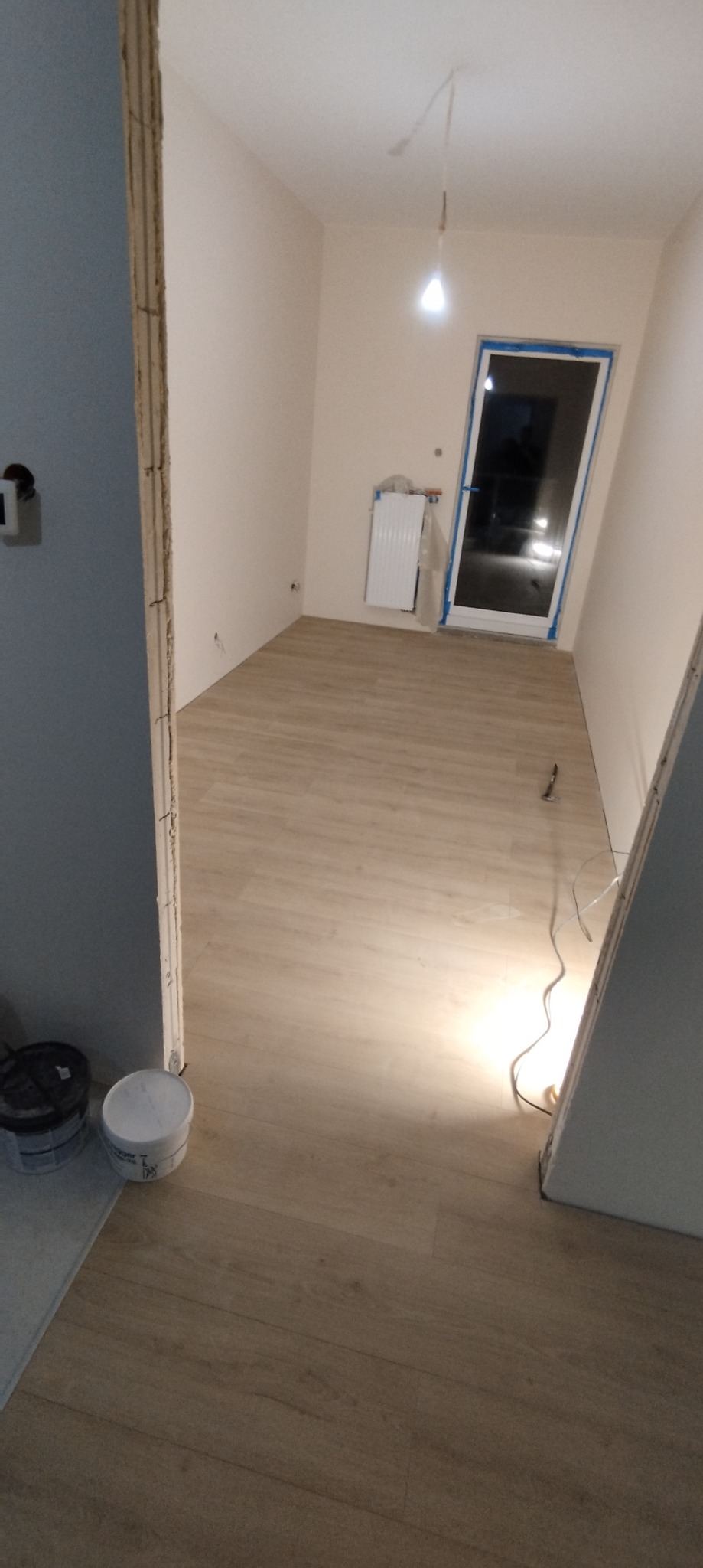 Wnętrze pokoju po remoncie, z nową podłogą imitującą drewno, świeżo pomalowanymi ścianami na jasny beż i niebiesko pomalowaną framugą drzwi. Grzejnik zabezpieczony folią. Widoczne puszki z farbą...