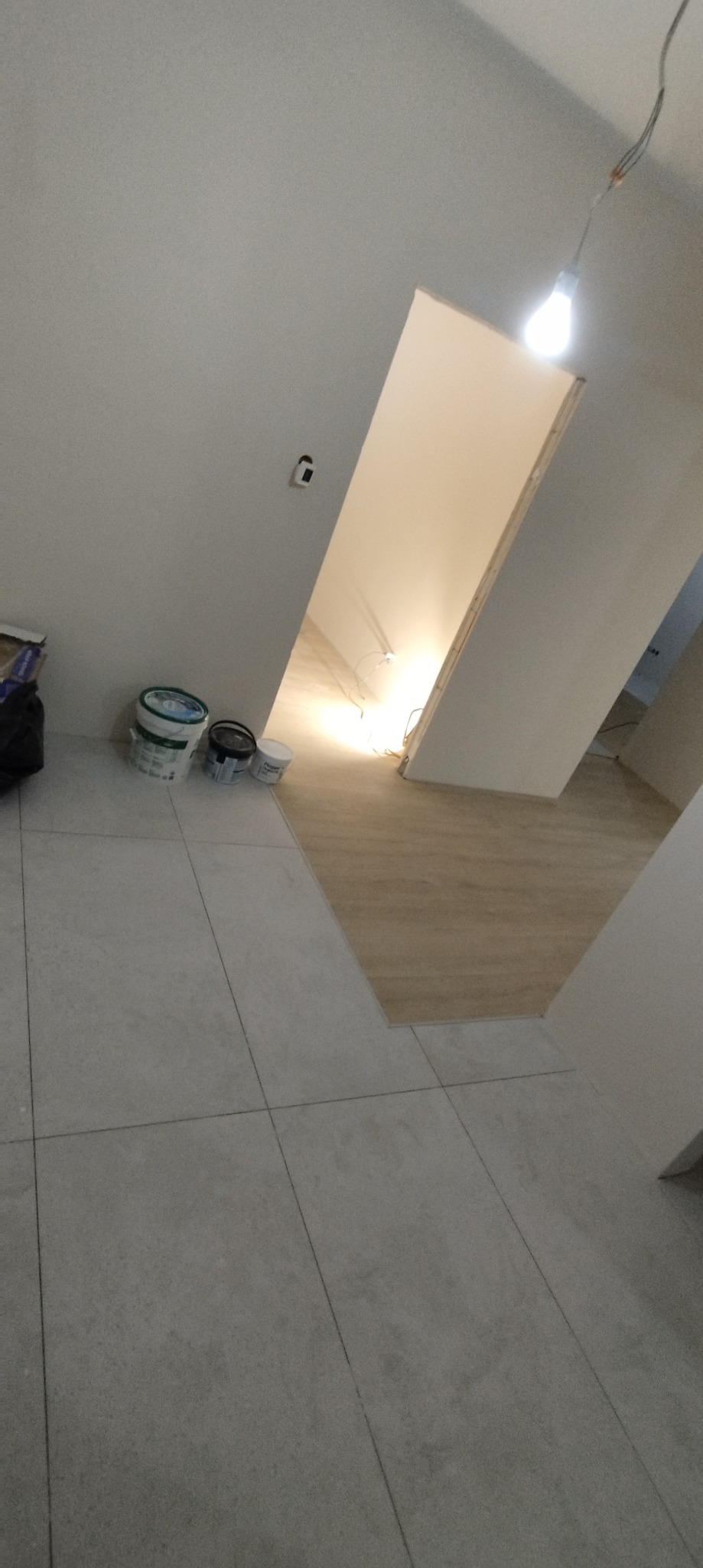 Wnętrze w trakcie remontu: widoczne puste wiadra po farbie przy ścianie, świeżo pomalowane ściany na biało, surowe oświetlenie żarówką, jasne płytki podłogowe i drewnopodobna podłoga w przejściu.