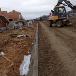 Budowa drogi dojazdowej: żółta koparka kołowa Mecalac MS w trakcie prac ziemnych, widoczne nowe krawężniki, studzienki kanalizacyjne i budynki w stanie surowym otwartym.