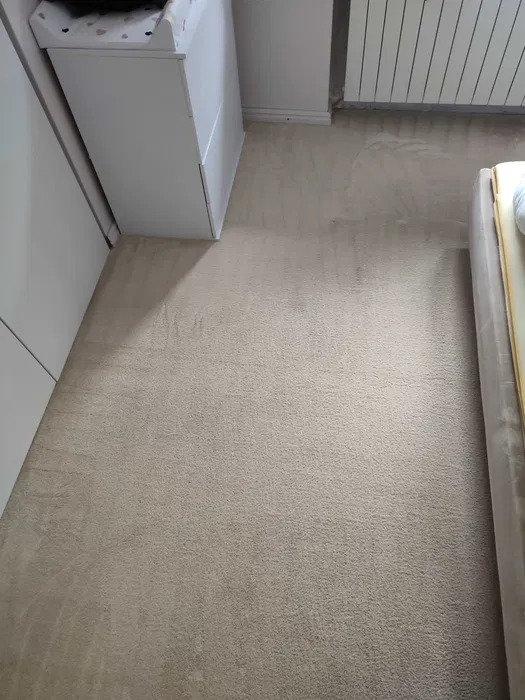 Beżowy dywan po czyszczeniu, widoczne odświeżone włókna, narożnik szafy, grzejnik i fragment łóżka w tle.