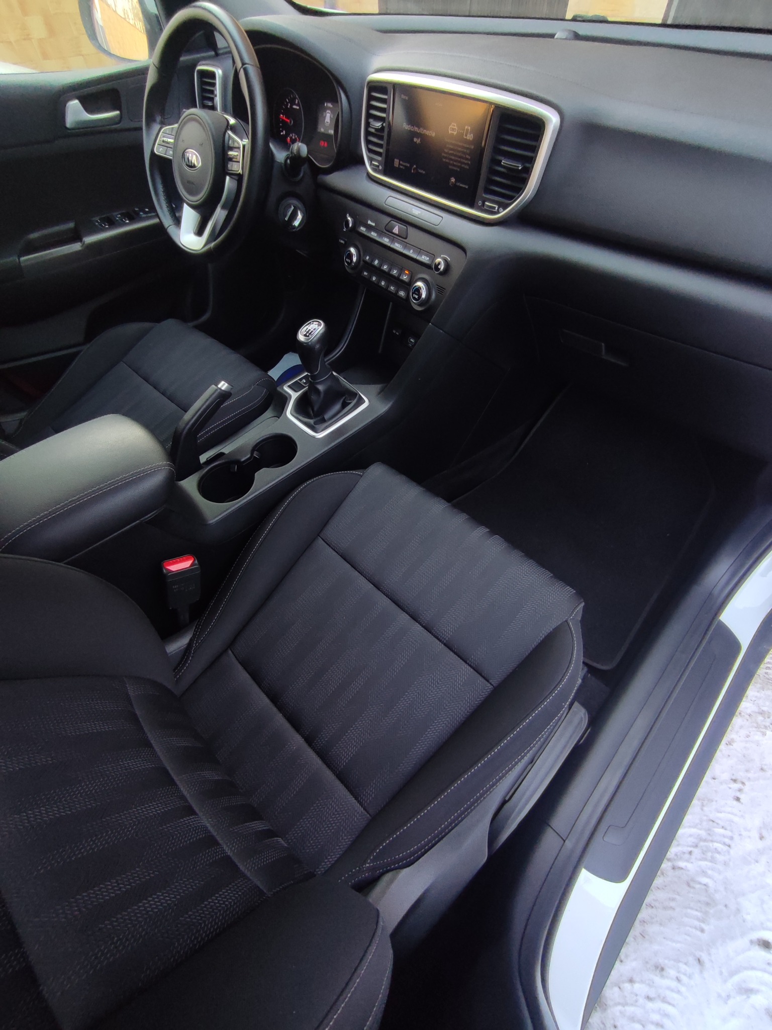 Wnętrze samochodu Kia Sportage z czarną tapicerką, widok z perspektywy fotela pasażera na deskę rozdzielczą, kierownicę z logo i dźwignię zmiany biegów.