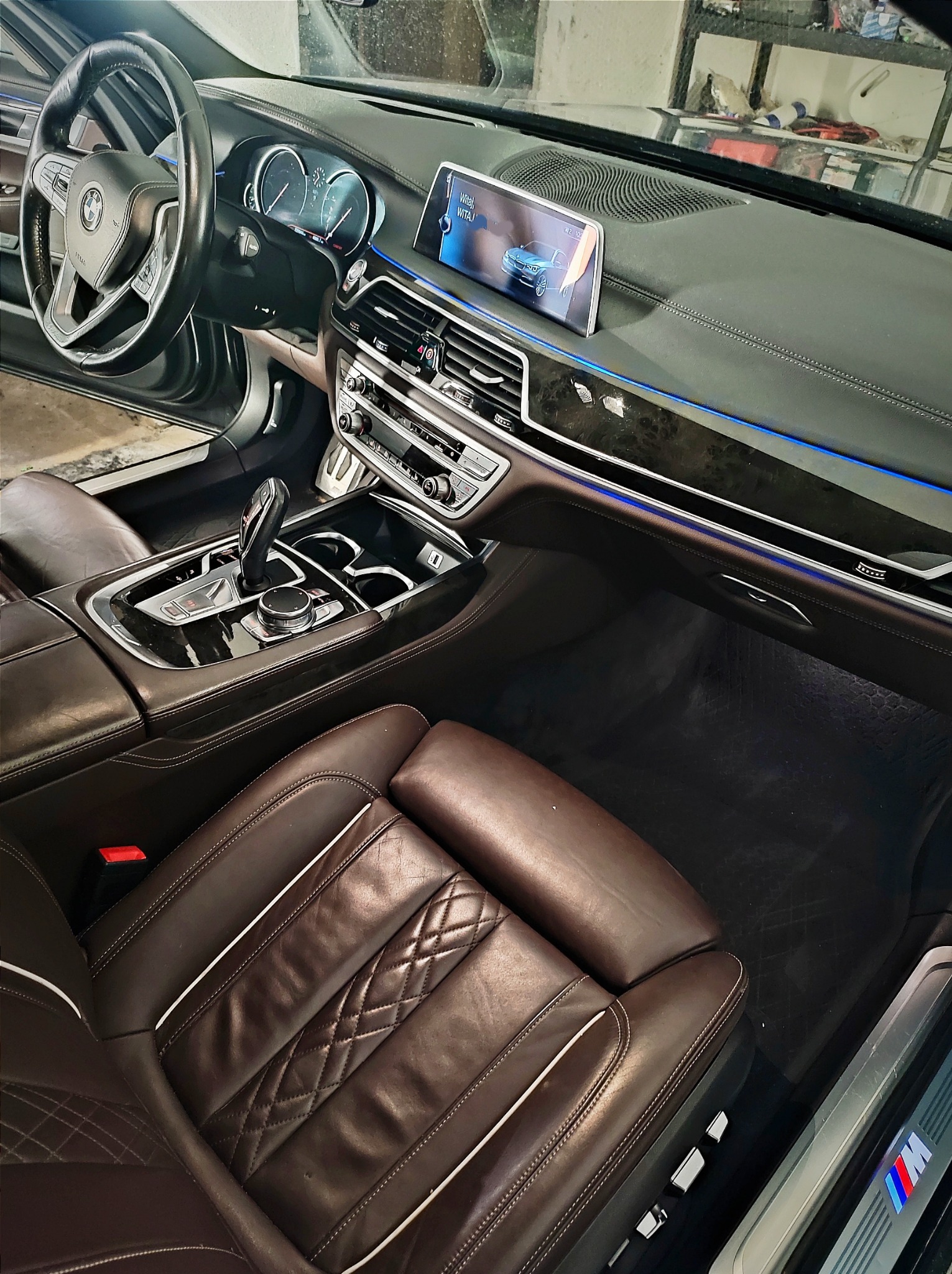 Wnętrze luksusowego samochodu BMW serii 7 z brązową skórzaną tapicerką, widoczne fotele z białymi przeszyciami, deska rozdzielcza z drewnianymi elementami i niebieskim podświetleniem, ekran...