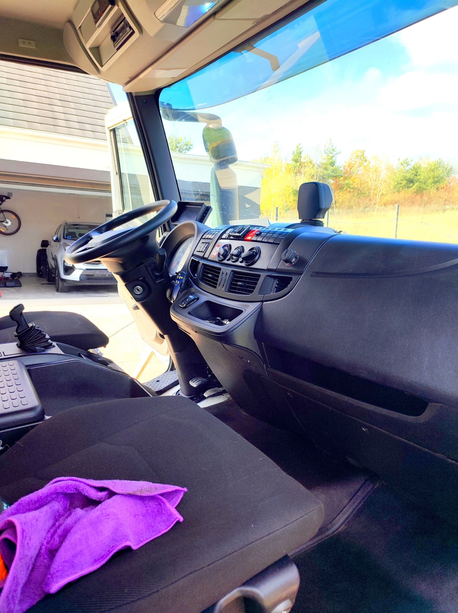Wnętrze kabiny samochodu ciężarowego z widoczną deską rozdzielczą, kierownicą i fotelem kierowcy z fioletową ściereczką, w tle garaż z zaparkowanym samochodem osobowym.