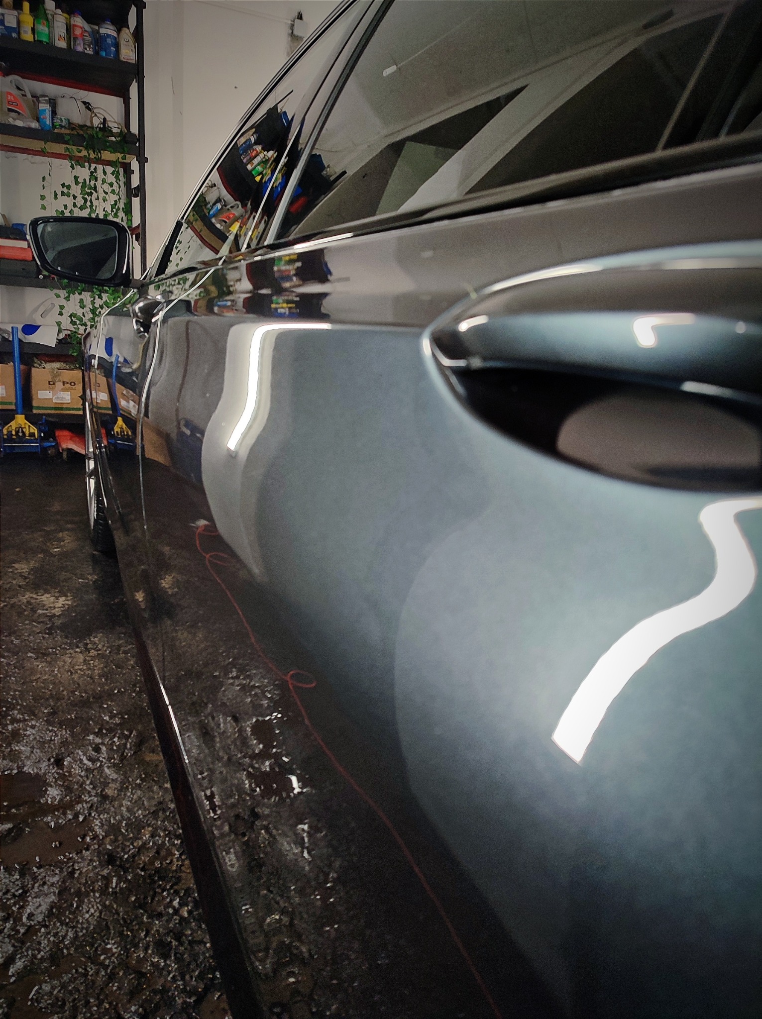 Błyszczący, szary bok samochodu z widocznym odbiciem światła, uchwytem drzwi i lusterkiem, stojący na mokrej posadzce w pomieszczeniu warsztatowym.