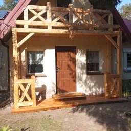 Ewsylwer zakład stolarski - Drewniany ganek z balustradą w surowym stanie przed domem jednorodzinnym z brązowymi drzwiami i schodami.