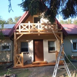 Ewsylwer zakład stolarski - Dom z nowo wybudowanym drewnianym tarasem i balkonem z ozdobnymi balustradami, drabina oparta o ścianę, widoczne panele słoneczne na dachu.