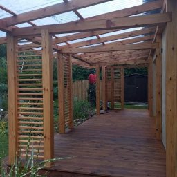 Ewsylwer zakład stolarski - Drewniana konstrukcja z zadaszeniem z przezroczystego tworzywa, tworząca zadaszone przejście, z drewnianą podłogą i ażurowymi ściankami, otoczona zielenią.