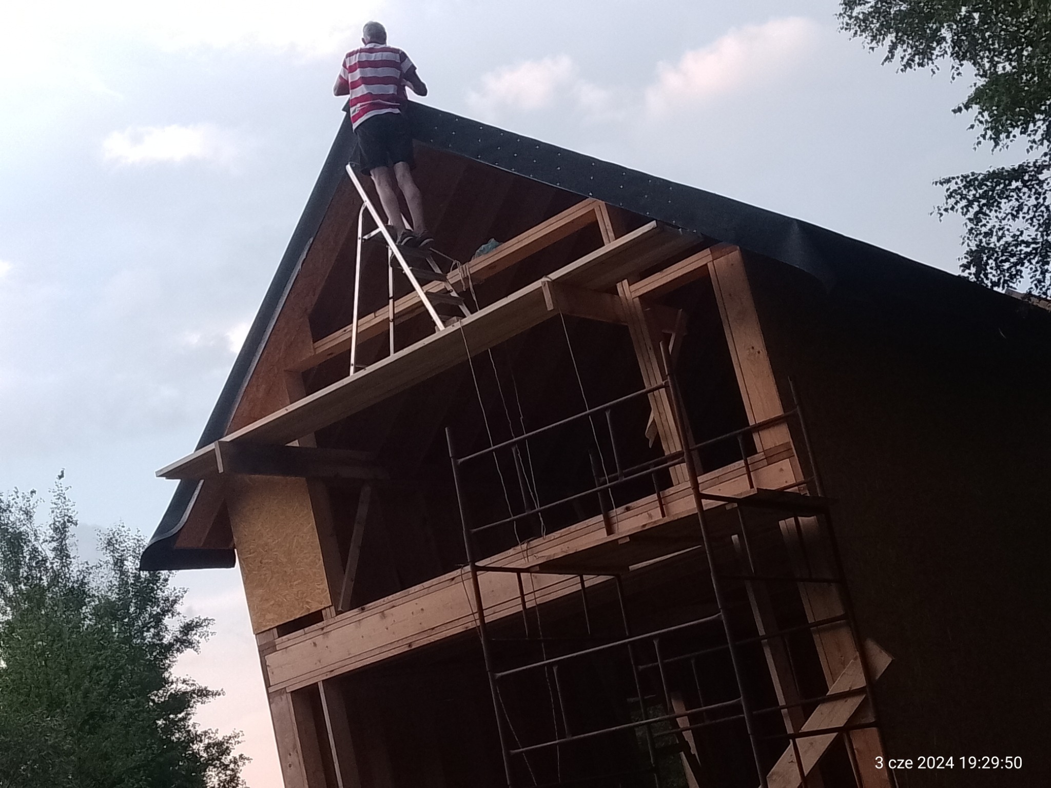 Montaż drewnianej konstrukcji dachu domu jednorodzinnego, widoczny mężczyzna na drabinie, drewniane elementy konstrukcyjne i rusztowanie.