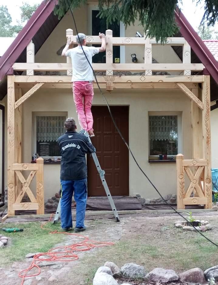 Dwóch mężczyzn montuje drewnianą konstrukcję werandy z dekoracyjnymi elementami, jeden na drabinie, drugi ją asekuruje. Widoczne narzędzia i przewody na trawniku.