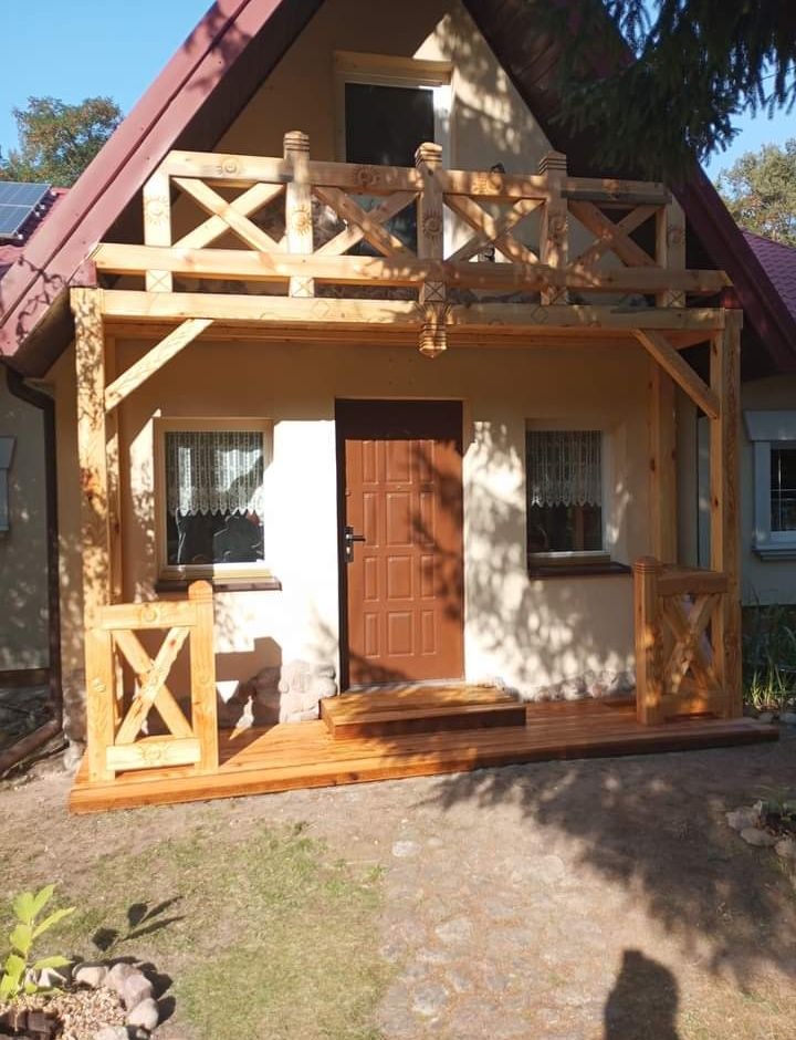 Drewniany ganek z balustradą w surowym stanie przed domem jednorodzinnym z brązowymi drzwiami i schodami.