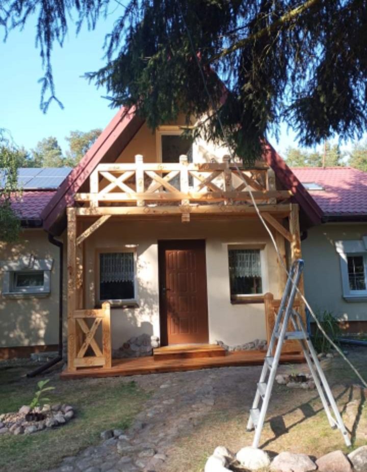 Dom z nowo wybudowanym drewnianym tarasem i balkonem z ozdobnymi balustradami, drabina oparta o ścianę, widoczne panele słoneczne na dachu.