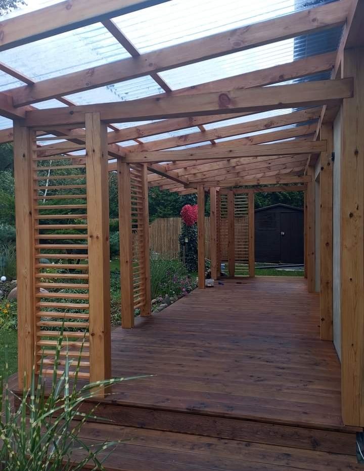 Drewniana konstrukcja z zadaszeniem z przezroczystego tworzywa, tworząca zadaszone przejście, z drewnianą podłogą i ażurowymi ściankami, otoczona zielenią.