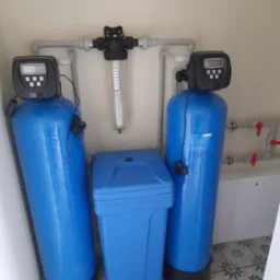 Kompletny system filtracji wody z dwoma niebieskimi kolumnami, filtrem pośrednim i zbiornikiem, zainstalowany w pomieszczeniu z wzorzystą podłogą.