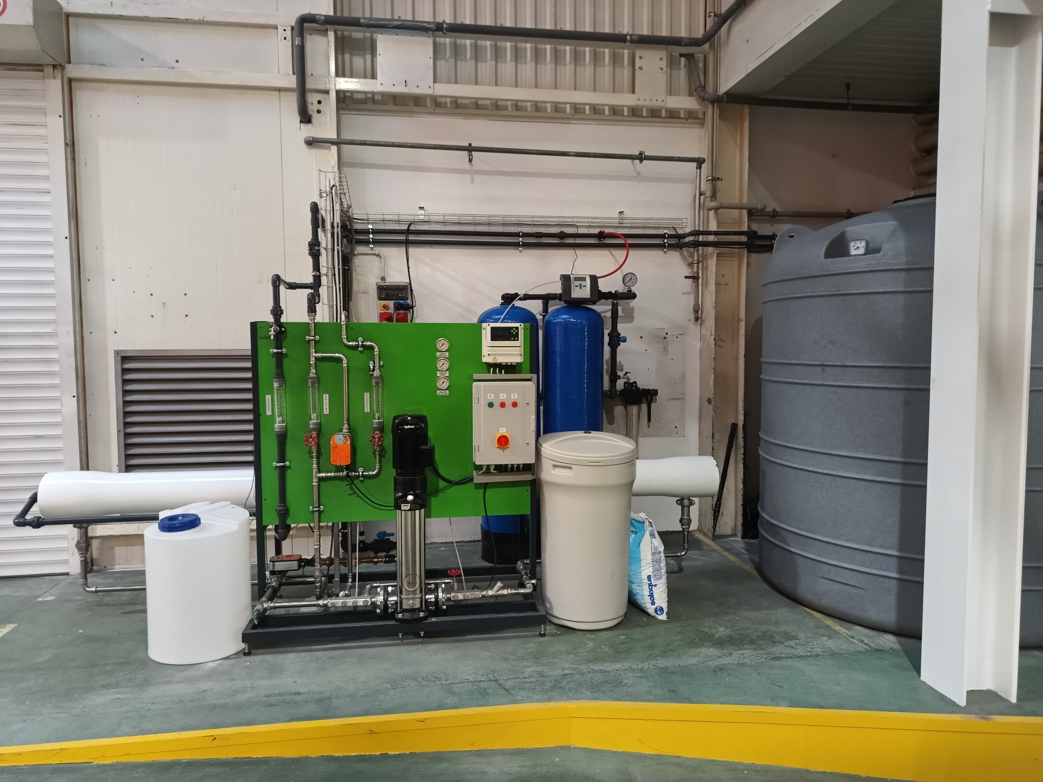 Kompletny system uzdatniania wody: filtr, zmiękczacz dwukolumnowy, odwrócona osmoza, zbiornik 3000l. Instalacja w hali, widoczne rury i zbiorniki.