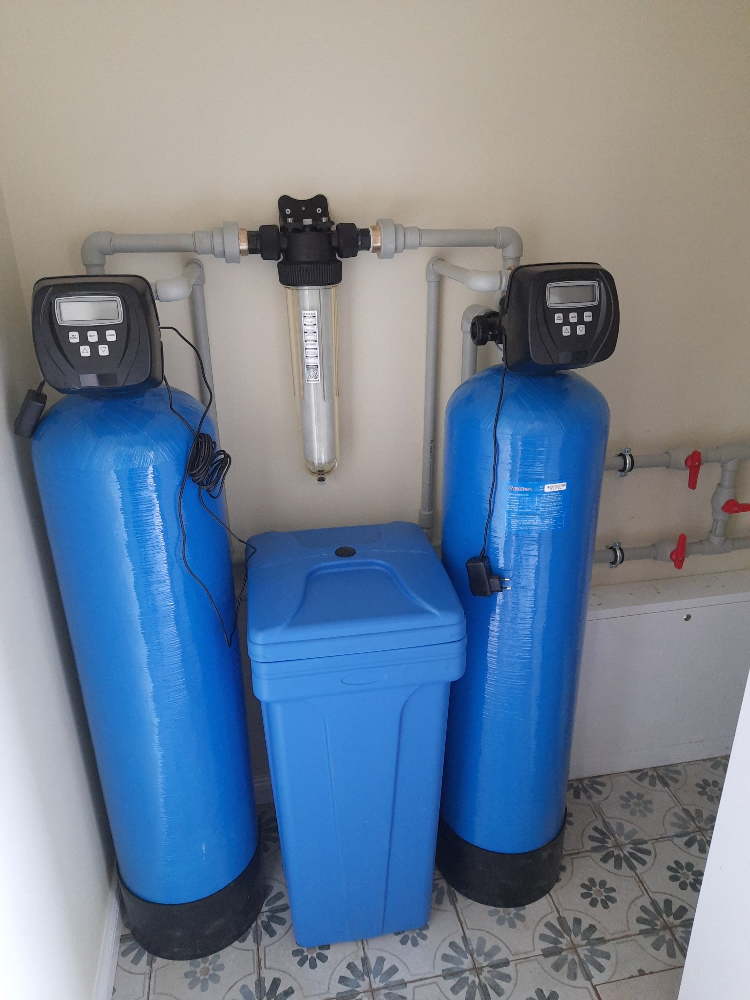 Kompletny system filtracji wody z dwoma niebieskimi kolumnami, filtrem pośrednim i zbiornikiem, zainstalowany w pomieszczeniu z wzorzystą podłogą.