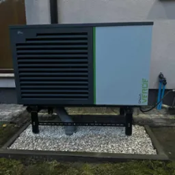 Zewnętrzna jednostka pompy ciepła Calla Verde na metalowej konstrukcji, z widoczną rurą odprowadzającą skropliny, otoczona ozdobnym żwirem na tle ściany budynku.