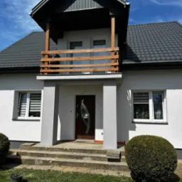 Nowoczesny dom murowany z białymi ścianami, szarą dachówką i drewnianą balustradą balkonu nad wejściem, widoczne schody wejściowe i starannie przycięte krzewy.