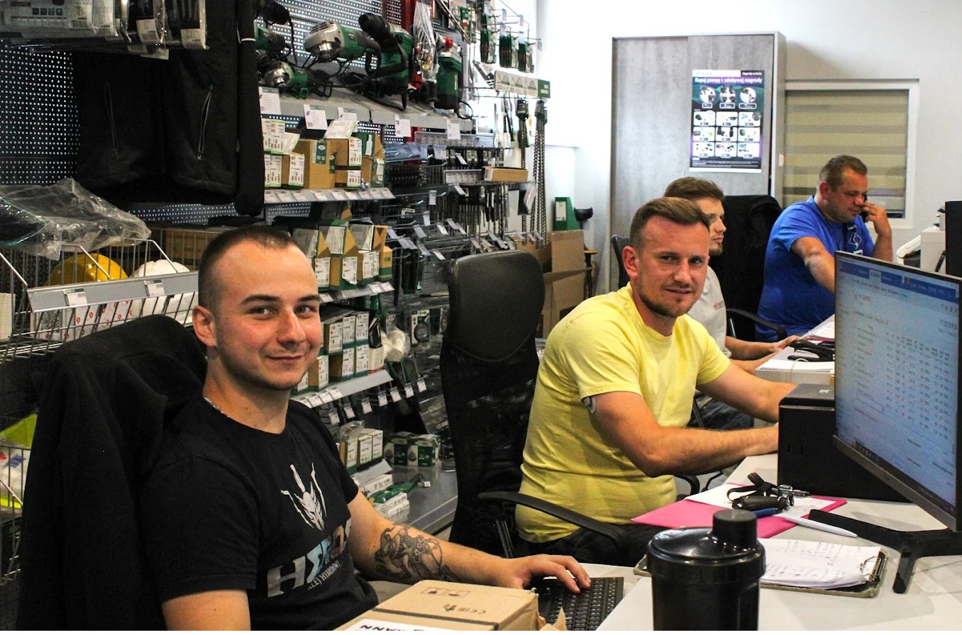 Zespół pracowników firmy budowlanej w biurze, z półkami pełnymi narzędzi i materiałów budowlanych w tle, pracujący przy komputerach.