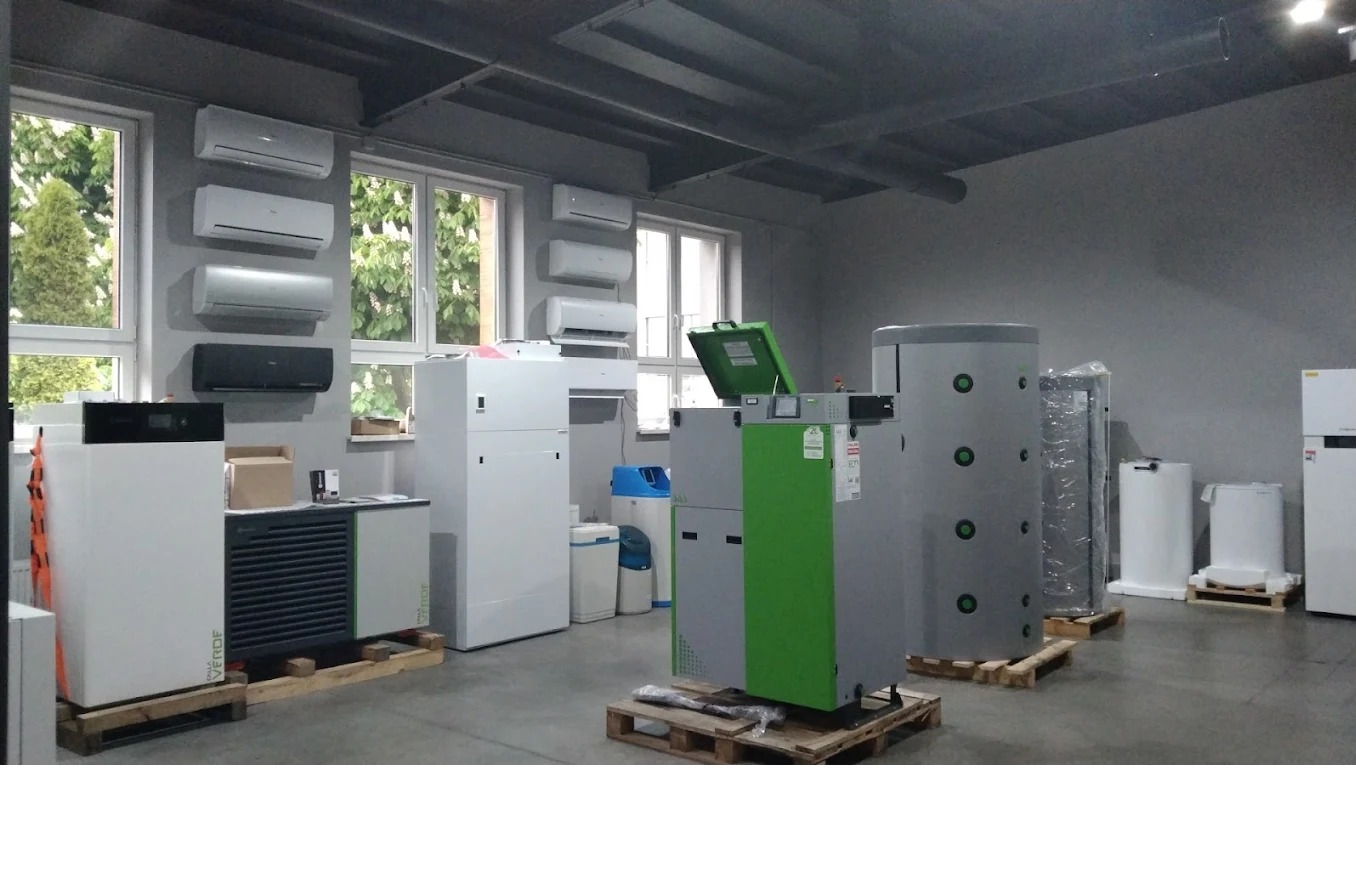 Wnętrze magazynu z wystawionymi kotłami, pompami ciepła i klimatyzatorami, część z nich umieszczona na drewnianych paletach.