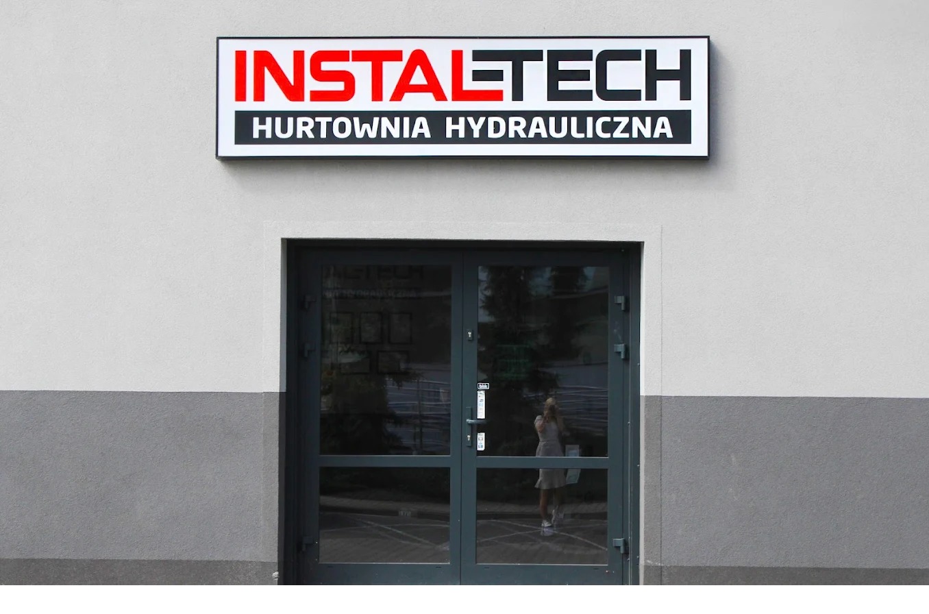 Fasada budynku hurtowni hydraulicznej Instaltech, z widocznym szyldem nad wejściem i odbiciem osoby w szklanych drzwiach.