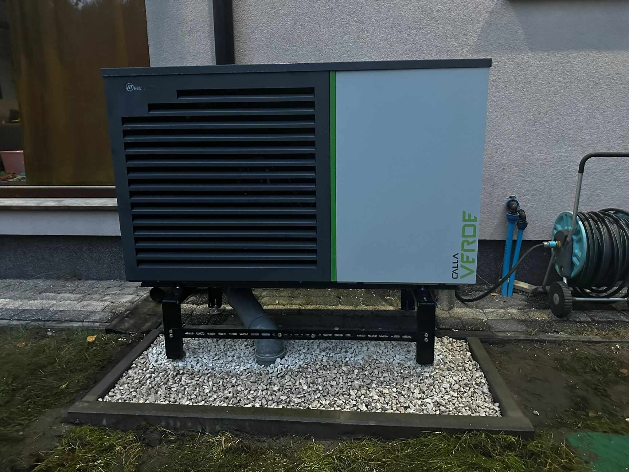 Zewnętrzna jednostka pompy ciepła Calla Verde na metalowej konstrukcji, z widoczną rurą odprowadzającą skropliny, otoczona ozdobnym żwirem na tle ściany budynku.