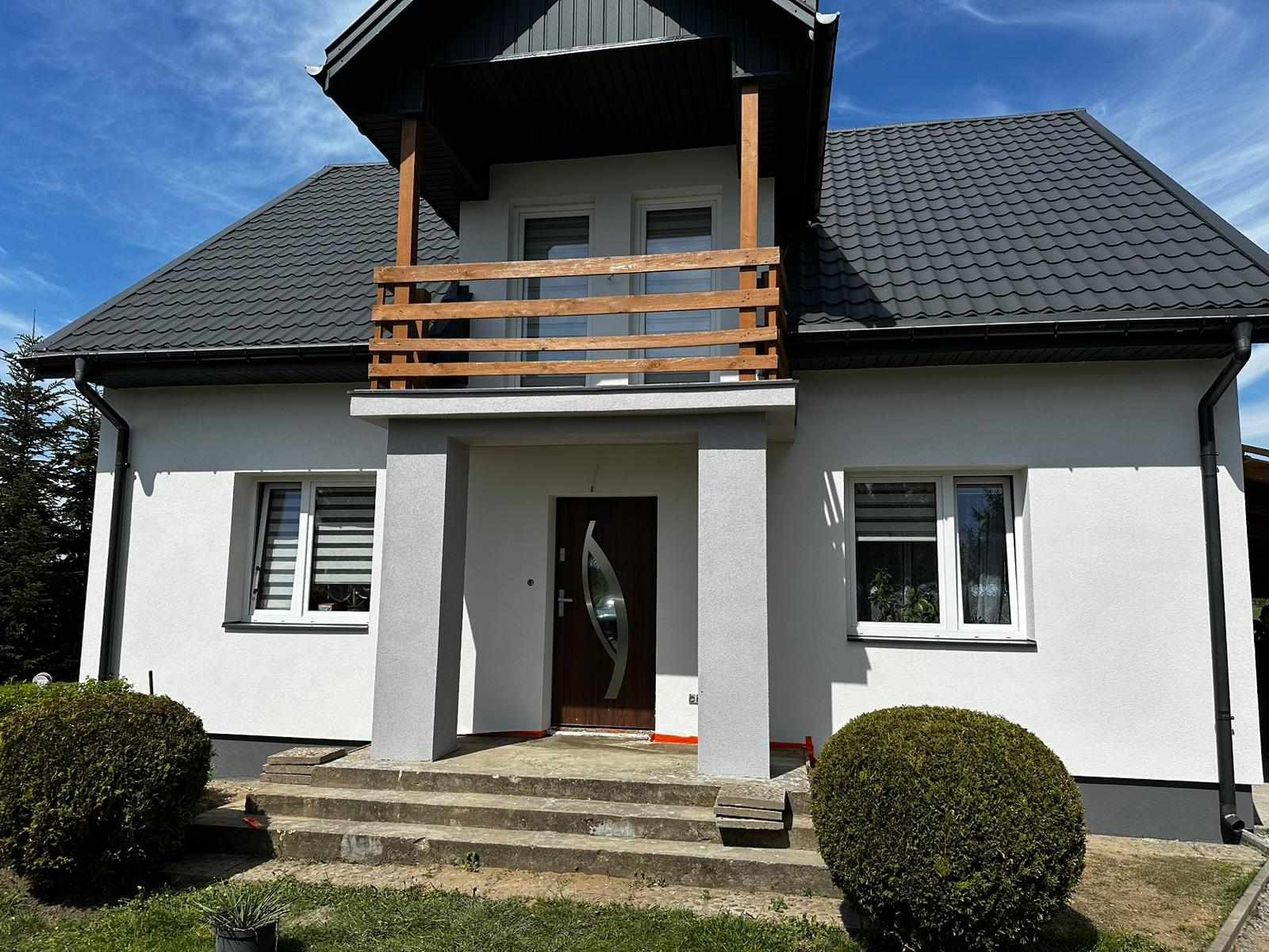 Nowoczesny dom murowany z białymi ścianami, szarą dachówką i drewnianą balustradą balkonu nad wejściem, widoczne schody wejściowe i starannie przycięte krzewy.