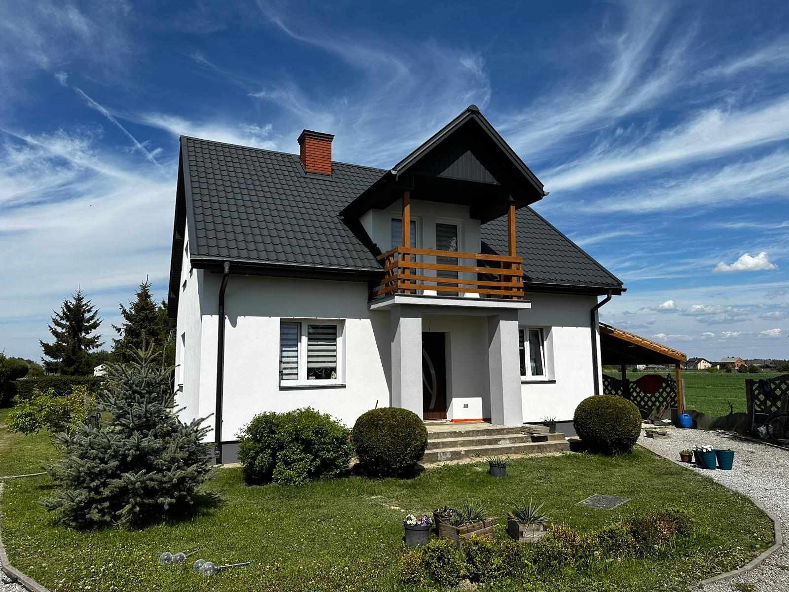 Gotowy dom murowany z balkonem, białą elewacją i ciemną dachówką na tle błękitnego nieba z chmurami.